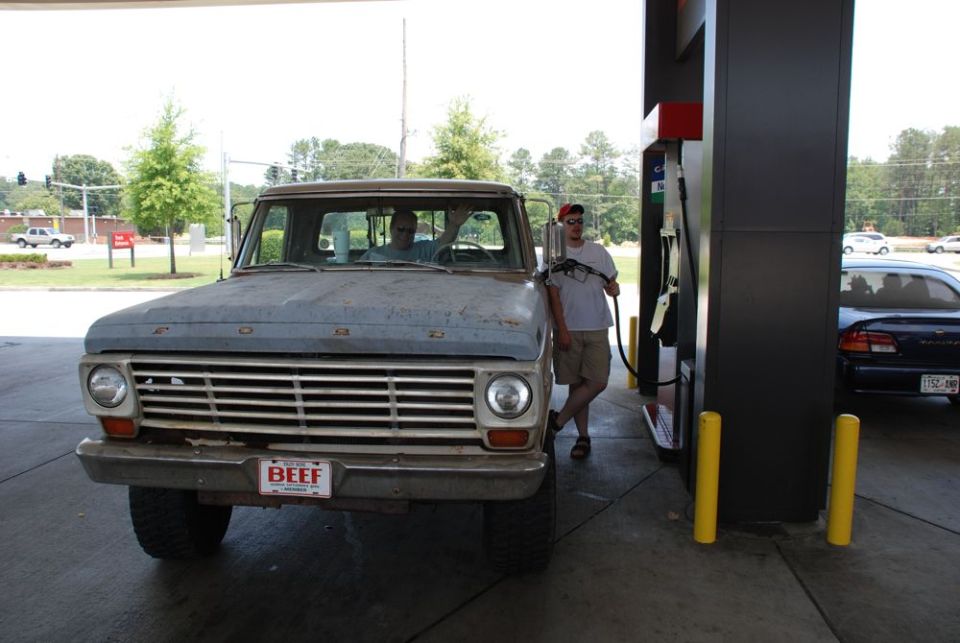 Front view of a weathered blue Ford pickup truck at a gas station with a person pumping gas