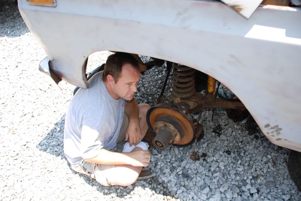 Man crouches under a light-colored vehicle's wheel well, examining the suspension and brake rotor