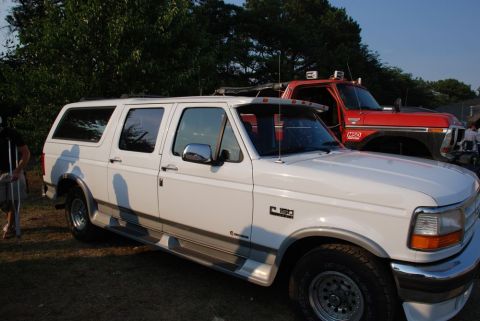 1996 Ford Bronco - A white 1996 Ford Bronco is parked next to a red truck, with a person on crutches visible on the left.