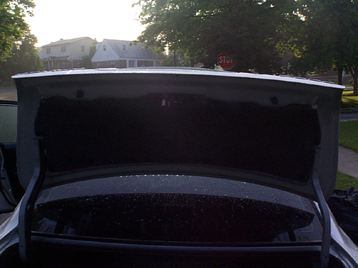 Open trunk lid of a light-colored sedan at dusk in a residential driveway near a stop sign