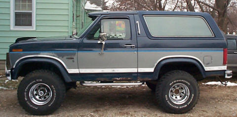 1986 Ford Bronco - Driver-side profile of a lifted two-tone blue-over-silver full-size Ford Bronco with a hardtop, parked on a dirt driveway beside a green ho…