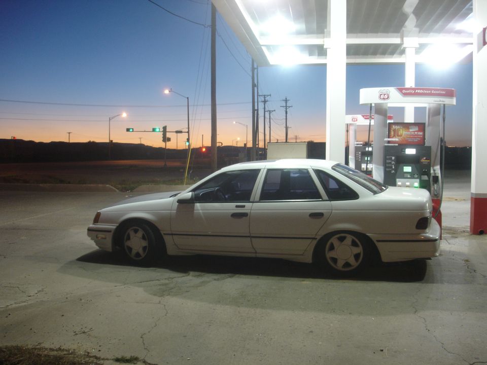 White four-door sedan parked at a gas station at dusk, side view.