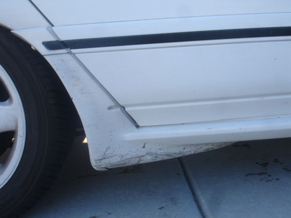 Close-up of damaged white car lower rear quarter panel with black trim.