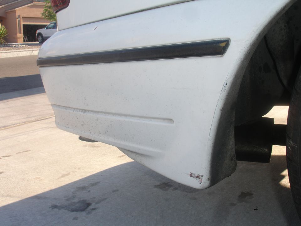 White sedan rear bumper corner with black trim and scuff marks, tire and exhaust visible.