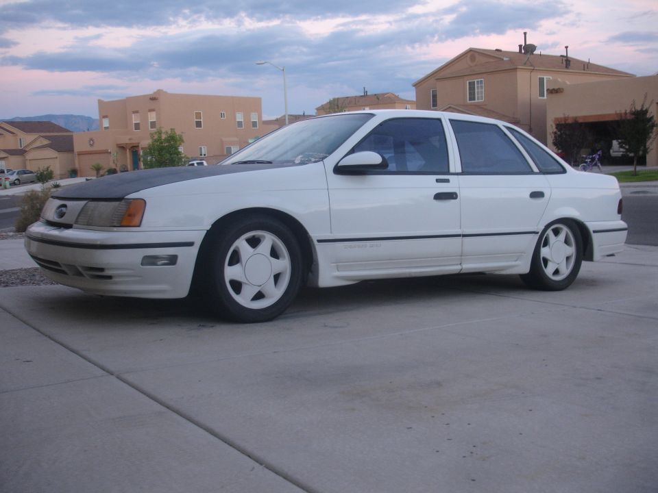 White 1990 Ford Taurus sedan with black hood wrap and aftermarket wheels parked on a driveway.