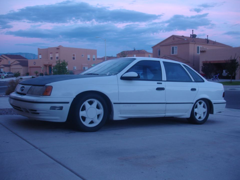 White 1990 Ford Taurus sedan with matte black hood and aftermarket wheels parked outside.