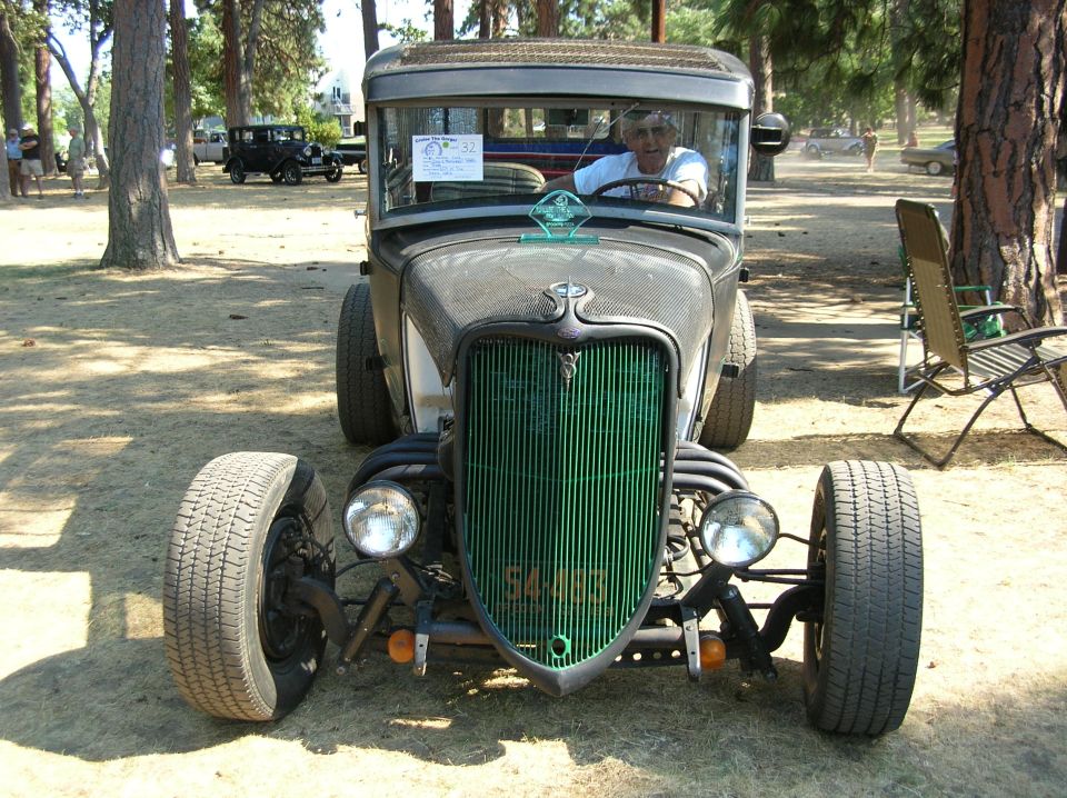 Front of a bare-metal hot rod with green grille and a man seated inside at a shaded car show