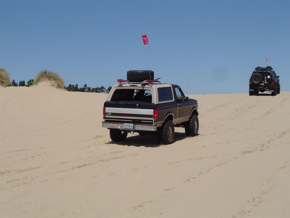 Two Ford Broncos driving on open sand dunes under blue sky