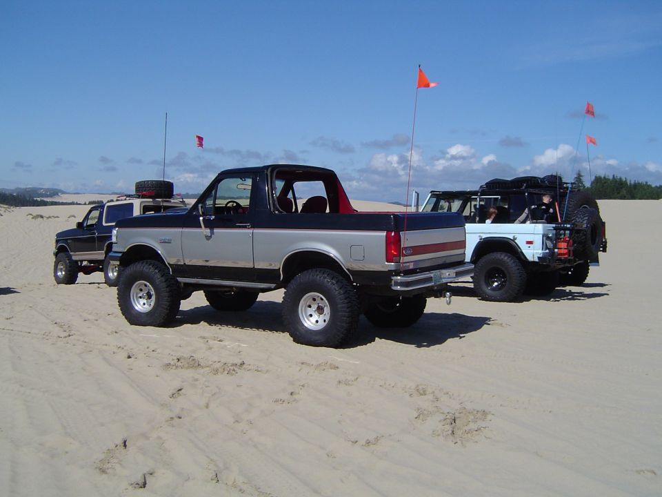 Several lifted Ford Broncos parked together on sand dunes with flags
