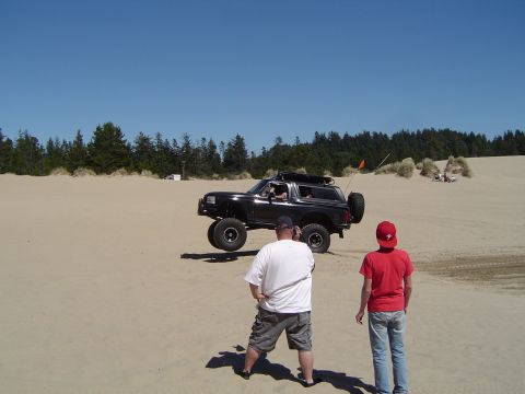 1989 Ford Bronco - A black 1989 Ford Bronco is captured mid-air as it jumps over sand dunes, with two onlookers observing the action.