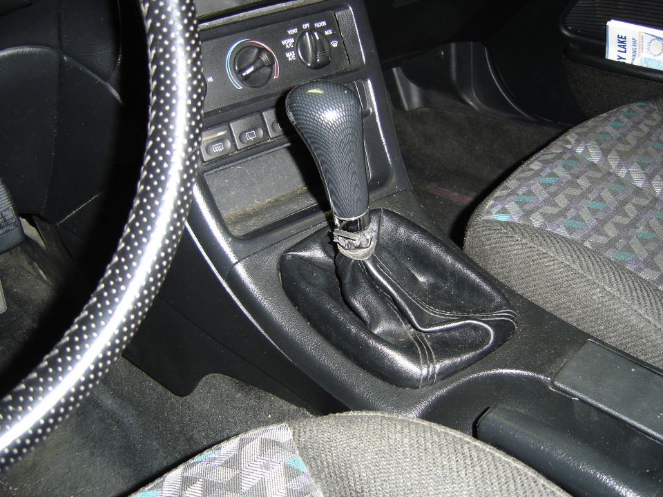 Car interior showing gear shifter, steering wheel, dashboard, and patterned seat.