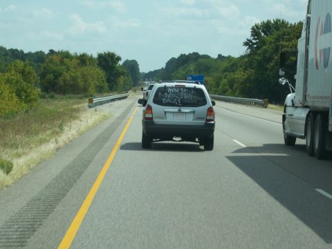 1996 Ford Bronco - The rear of a silver SUV is visible on a highway, with text written on its rear window that reads "NEW SHICK OVER, STAY BACK!". A large whi…