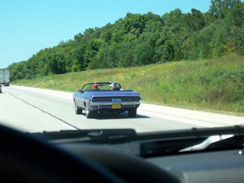 1996 Ford Bronco - A purple convertible muscle car is driving on a highway with two people inside. The car has a yellow license plate that reads "PP0000".