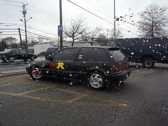 Black Honda Civic hatchback with red and yellow R decals, rear 3/4 in rainy lot