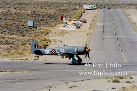 1992 Ford Bronco - A vintage gray Hawker Sea Fury airplane with the markings "BC-OL 114" taxis on a runway. Several other small aircraft are parked in the bac…