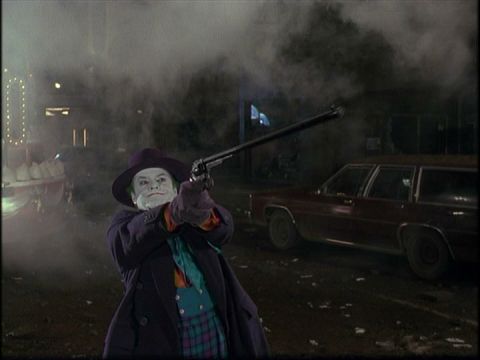 1988 Mercury Grand Marquis - A man dressed as the Joker aims a revolver forward in a smoky, dimly lit outdoor scene. A maroon station wagon is parked behind him on a di…