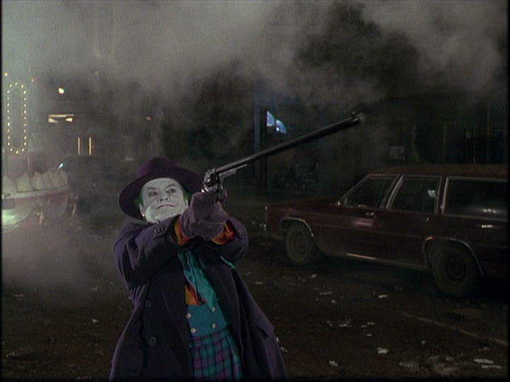The Joker aims a gun in front of a maroon station wagon.