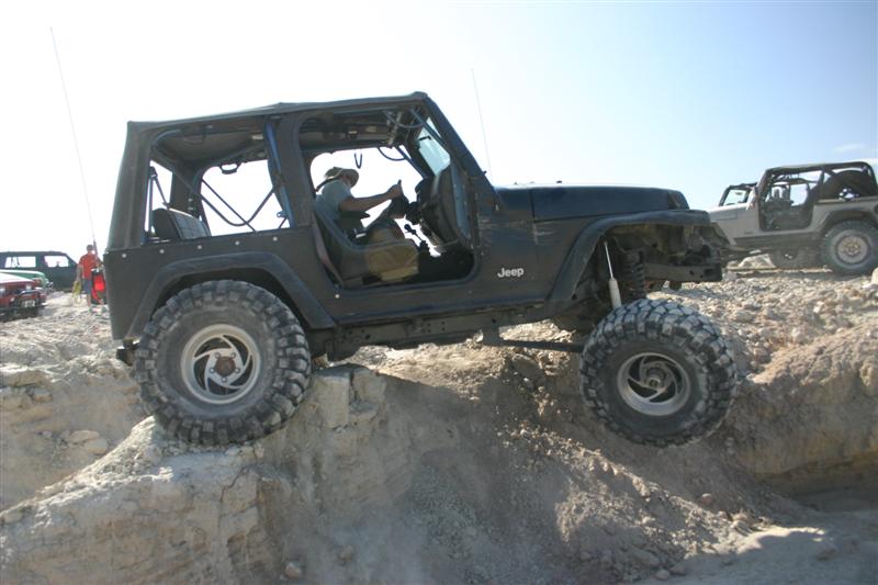 Black Jeep TJ off-roading, front passenger tire lifted over rocky terrain.