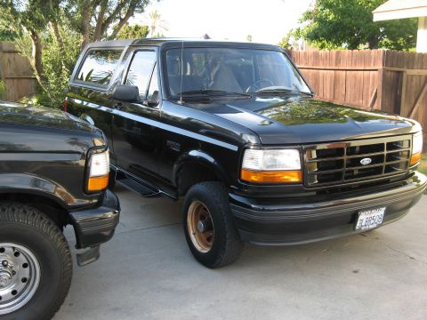 1986 Ford Bronco - A black 1980s Ford Bronco is parked in a driveway next to another black vehicle. The Bronco has a chrome grille, orange turn signals, and a…