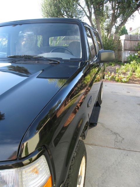 1986 Ford Bronco - A black Ford Bronco is parked on a concrete driveway with a wooden fence and garden in the background. The vehicle is viewed from a low ang…