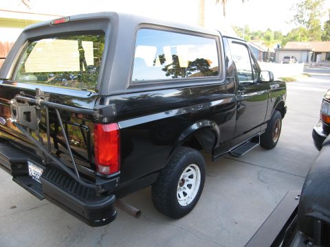 1986 Ford Bronco - The rear driver's side of a black Ford Bronco is shown parked on a concrete driveway. Visible modifications include a spare tire carrier mo…