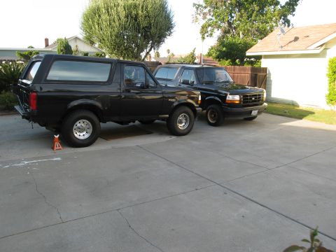 1986 Ford Bronco - Two black Ford Broncos are parked in a driveway. The Bronco on the left is supported by an orange jack stand under its rear axle.