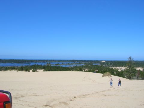 1996 Ford Bronco - A red vehicle is partially visible in the lower left corner of the frame, parked on a sandy dune overlooking a body of water and a forest.…