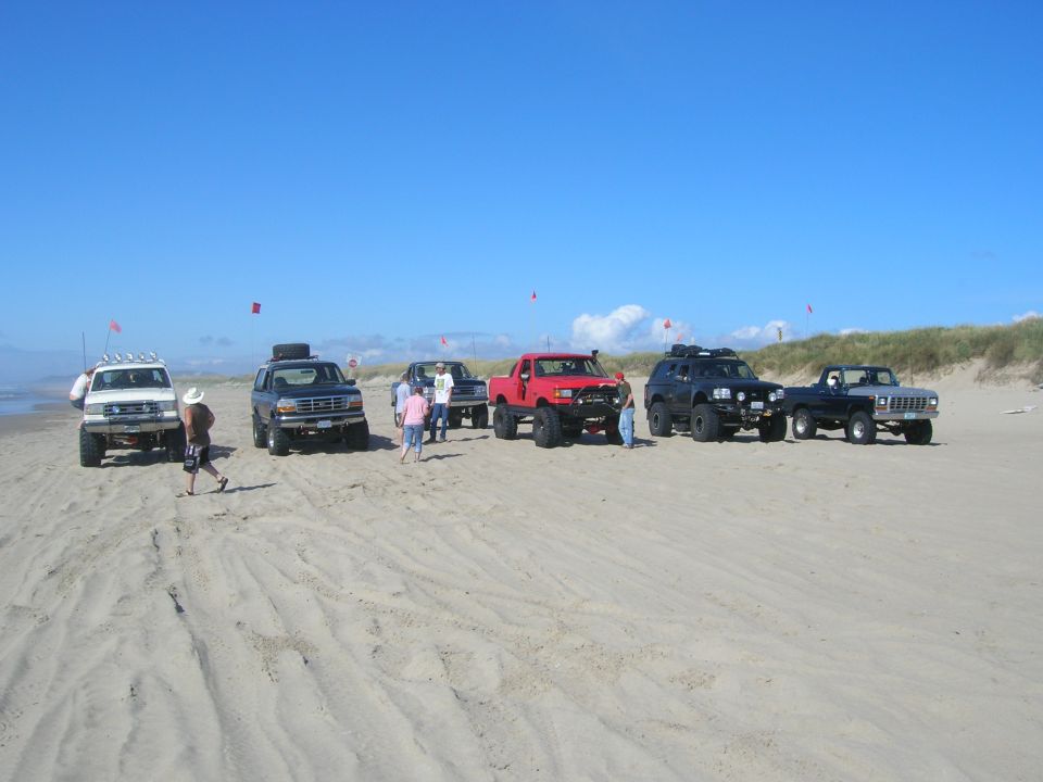 Off-road trucks and SUVs parked on a sandy beach with people around.
