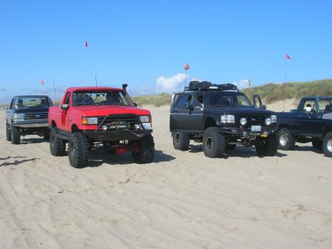 1996 Ford Bronco - Several modified off-road trucks and SUVs are parked on a sandy beach under a clear blue sky. A red Ford F-150 pickup is in the foreground,…