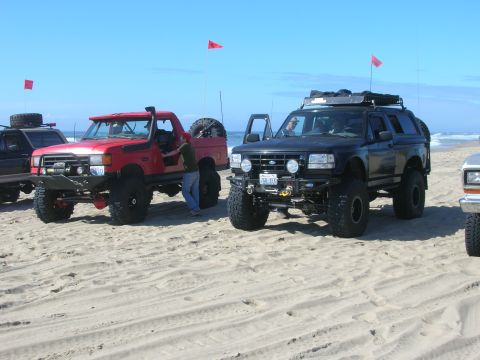 1996 Ford Bronco - Two lifted off-road trucks, a red Ford pickup and a black Ford Bronco, are parked on a sandy beach with the ocean in the background. Both v…
