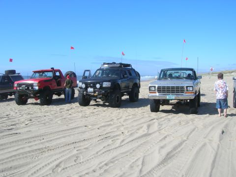 1996 Ford Bronco - A line of off-road vehicles are parked on a sandy beach under a clear blue sky, with red flags planted in the sand.