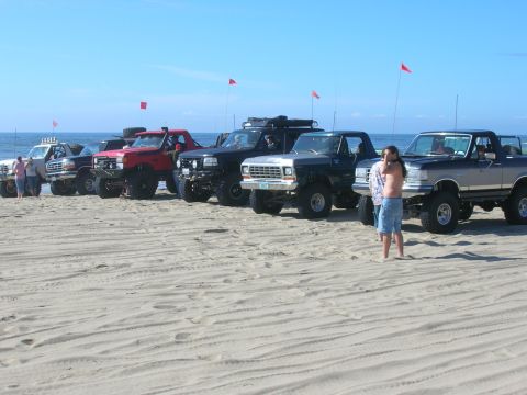 1996 Ford Bronco - Several lifted Ford trucks and SUVs are parked on a sandy beach with the ocean in the background under a clear blue sky. A man in blue shor…