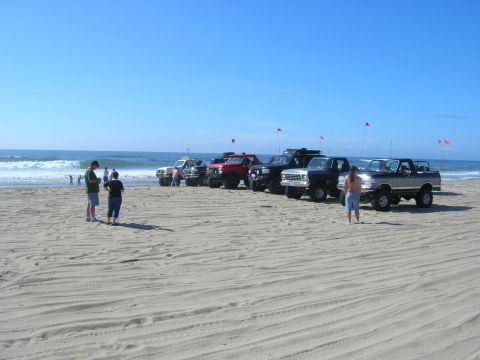 1996 Ford Bronco - Several lifted pickup trucks and SUVs are parked on a sandy beach with the ocean in the background under a clear blue sky. A few people are…