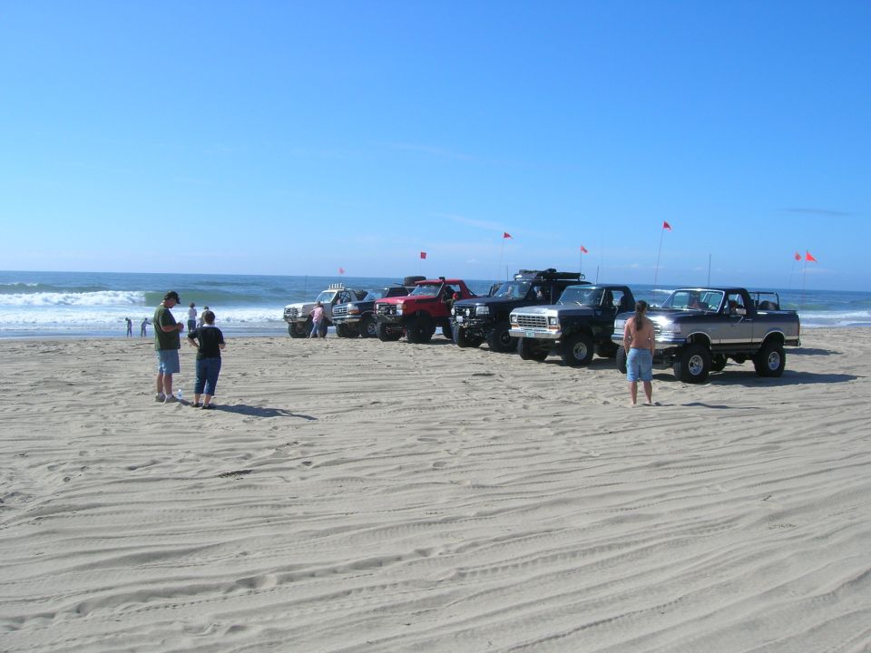 Several lifted pickup trucks and SUVs parked on a sandy beach with the ocean and blue sky.