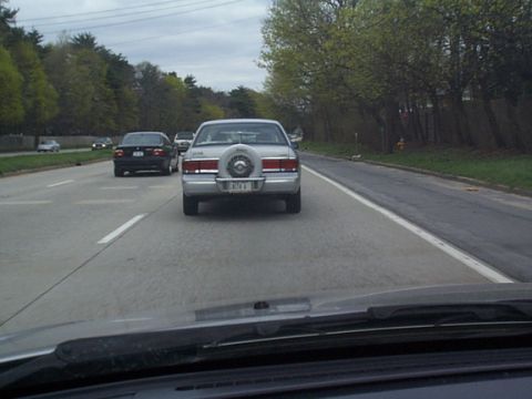 2002 Nissan Altima - A sedan with a spare tire mounted on its trunk drives on a highway, with another car in front.