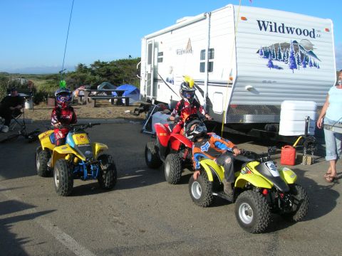 1996 Ford Bronco - Three children in riding gear are positioned on yellow and red ATVs in front of a Wildwood travel trailer. The scene appears to be a campgr…