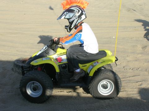1996 Ford Bronco - A child wearing a helmet with an orange mohawk rides a yellow Suzuki quad bike on a sandy surface. The quad bike has black fenders and tire…