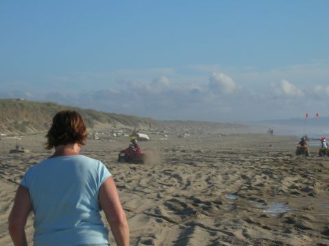 1996 Ford Bronco - A woman in a light blue t-shirt stands on a sandy beach, facing away from the camera. In the distance, several people are riding ATVs on th…