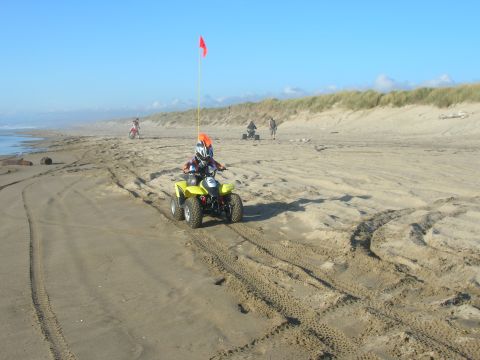 1996 Ford Bronco - A child wearing a helmet rides a yellow ATV with a red flag on a sandy beach, with other ATVs and people in the background.
