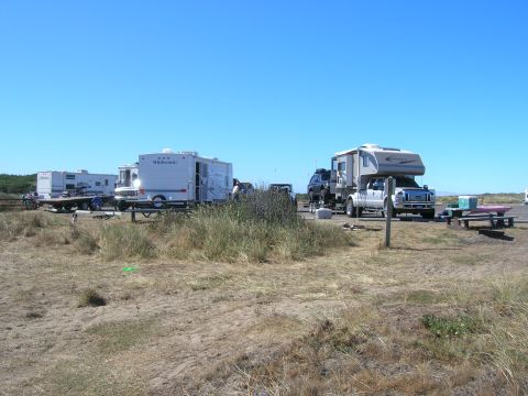 1996 Ford Bronco - A pickup truck with a camper shell is parked next to a picnic table and a pink kayak on a sandy area with sparse vegetation.