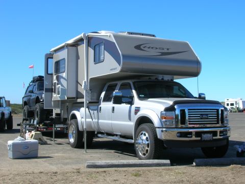 1996 Ford Bronco - A silver Ford F-series truck with a Host camper shell is parked on a dirt lot next to a trailer carrying a black SUV. The truck has a chrom…