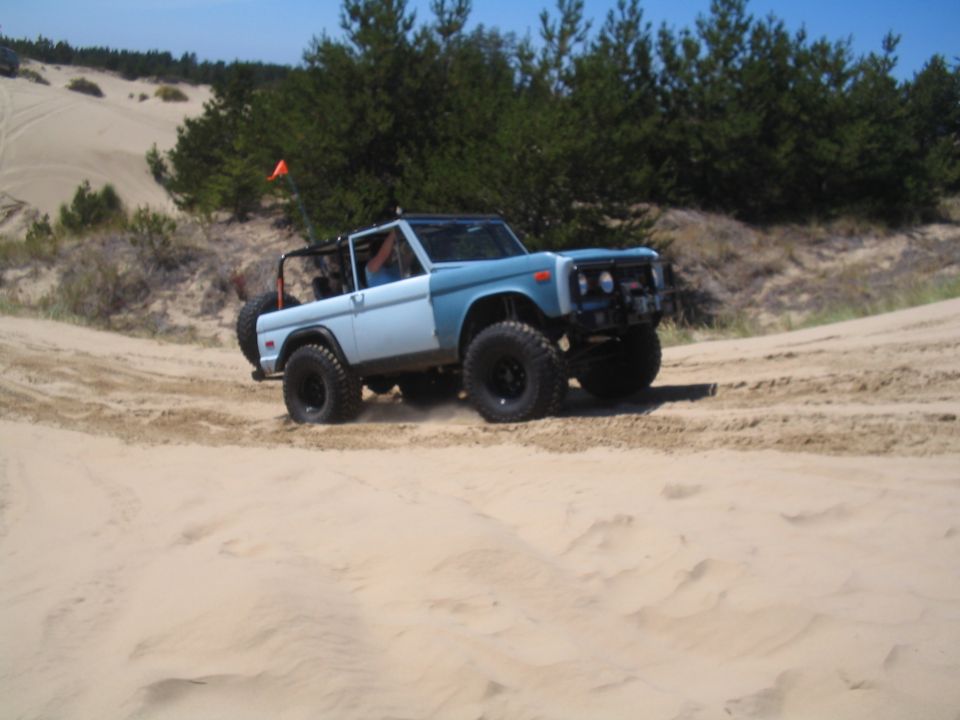 Light blue and white Ford Bronco with off-road tires driving on sand dunes.