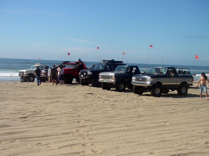 Lifted trucks and SUVs on a beach with people and the ocean.