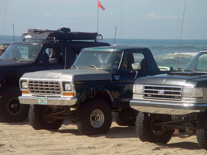 Three lifted Ford trucks parked on a sandy beach, with the ocean in the background. The middle truck is a dark green Bronco.