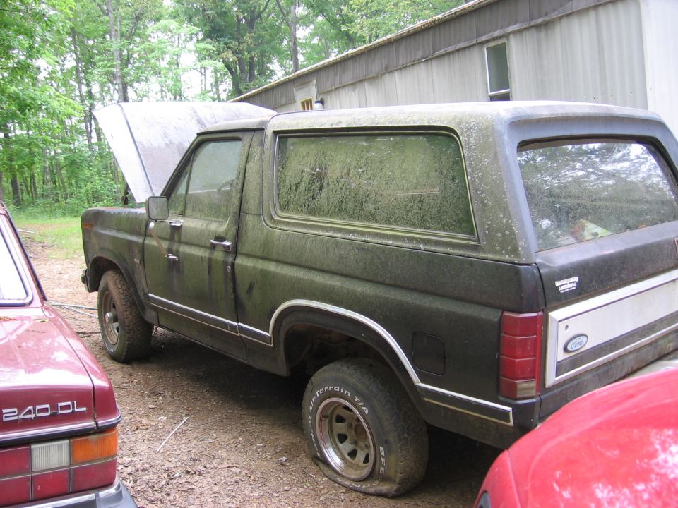 Moss-covered dark Ford Bronco with hood up, parked on dirt next to a metal building and a red Volvo 240 DL.