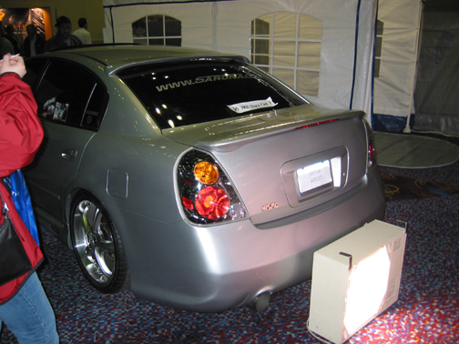 Silver Nissan Altima sedan at indoor car show, rear 3/4, chrome wheels, altezza tails