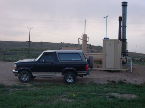 1996 Ford Bronco - A black 1996 Ford Bronco is parked on a dirt road near industrial equipment under an overcast sky.