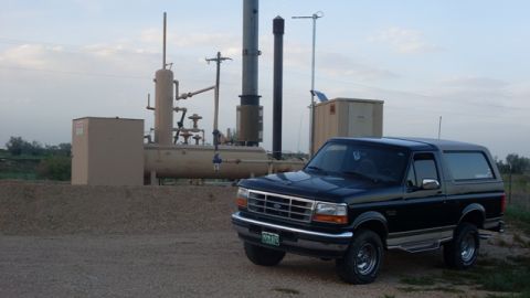 1996 Ford Bronco - A black 1996 Ford Bronco is seen parked in front of industrial processing equipment under a cloudy sky.