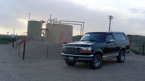 1996 Ford Bronco - A black 1996 Ford Bronco is parked in front of large industrial storage tanks under a cloudy sky.