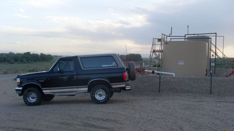 1996 Ford Bronco - A black 1996 Ford Bronco is parked on a gravel lot next to large industrial storage tanks under a cloudy sky.